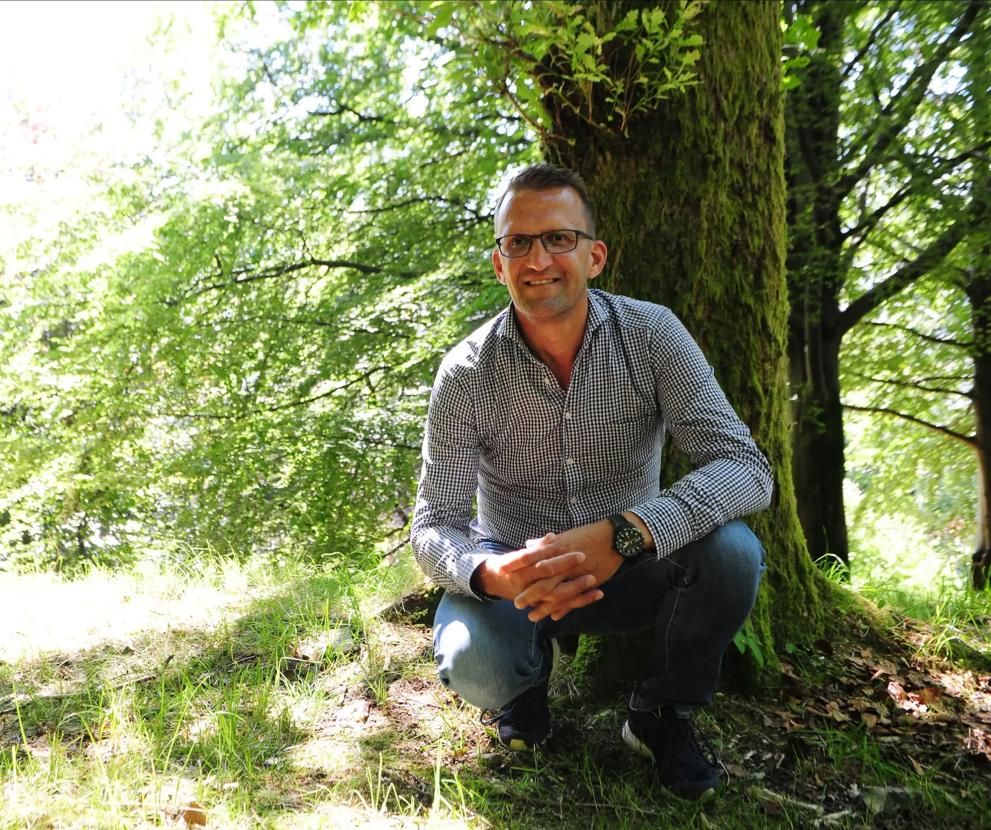 Ove sitande på huk framfor eit tre. Foto.